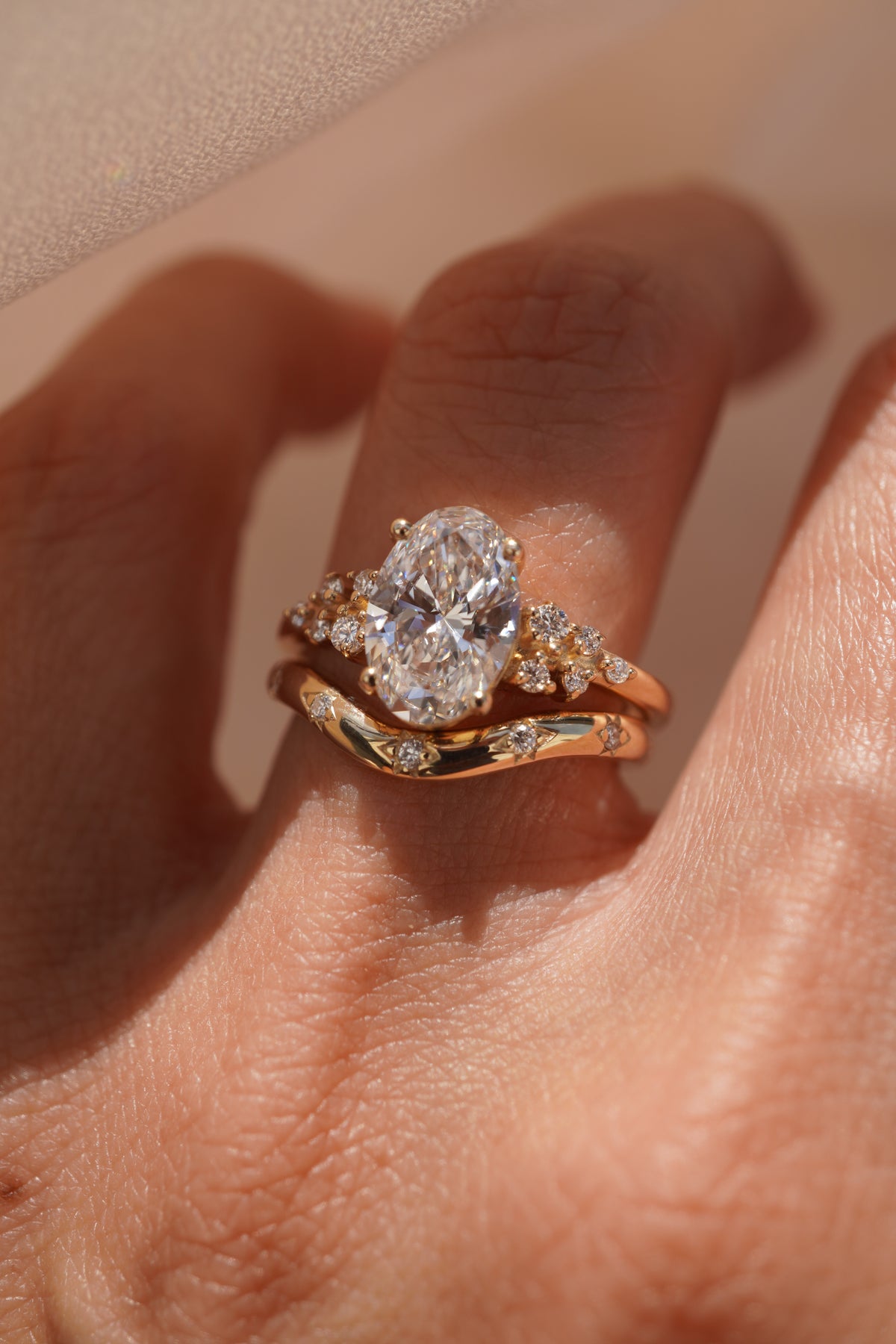 A close-up of a hand wearing a gold engagement ring with an oval-cut center stone and smaller accent stones, with a matching gold band partially visible underneath.